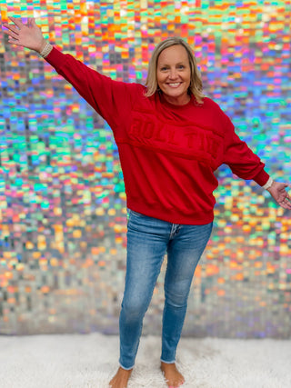 A smiling woman with blonde hair stands barefoot on a white rug, wearing a red crewneck sweatshirt with 'ROLL THE' in raised letters across the chest and blue skinny jeans. She is posing with one arm raised in front of a colorful, reflective mosaic tile wall.
