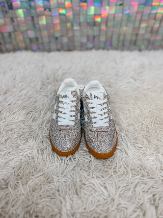 A pair of low-top sneakers covered in silver rhinestones with white laces and gum soles, displayed on a white shag rug in front of a holographic tile wall.
