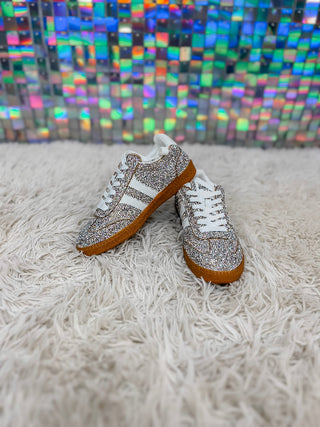 A pair of low-top sneakers covered in silver rhinestones, featuring white laces, white side stripes, and brown gum soles, resting on a white shag rug in front of a colorful, iridescent mosaic wall.