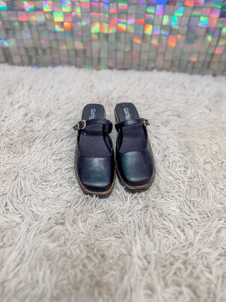 A pair of dark navy blue leather clog-style mule sandals with a single strap and a small gold buckle, resting on a white shag rug in front of a holographic tile wall.