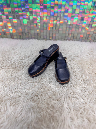 A pair of dark navy blue Mary Jane clogs with brown stitching and a small gold buckle, resting on a white shag rug in front of a colorful mosaic tile wall.