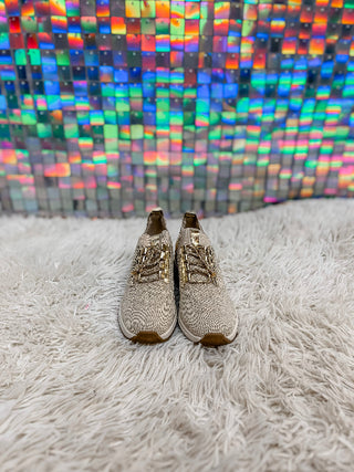 A pair of beige knit lace-up sneakers with gold-tone metal eyelets and brown soles, displayed on a white shag rug in front of a colorful holographic tile wall.