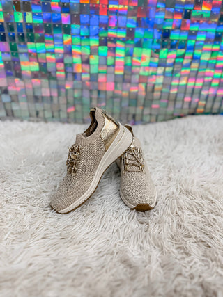 A pair of beige knit lace-up sneakers with metallic gold accents, displayed on a white shag rug in front of a colorful holographic tile wall.