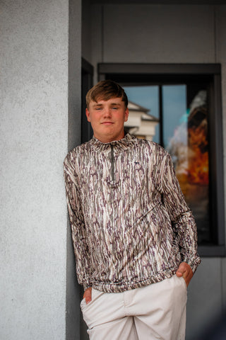 A young man wearing a high-neck, long-sleeve zip-up jacket with an abstract black, white, and gray brushstroke pattern, paired with off-white trousers, standing against a gray concrete pillar.