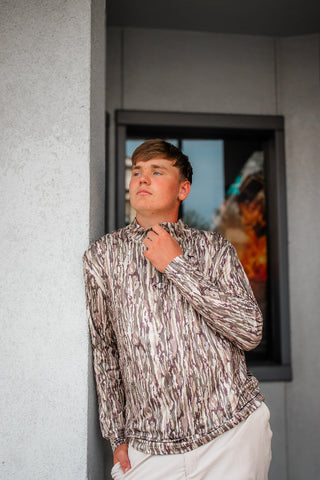 A young man wearing a brown and beige abstract patterned long-sleeve shirt and white pants, standing in front of a gray textured wall and a dark-framed window.