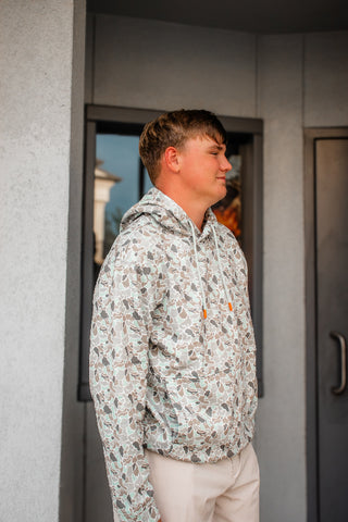 A man wearing a light beige hoodie with an all-over brown and gray paisley pattern, paired with beige pants, standing in profile in front of a gray building wall.
