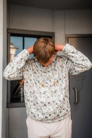 A man wearing a light beige hooded sweatshirt with a brown and gray abstract pattern, standing with his hands behind his head in front of a gray building wall.