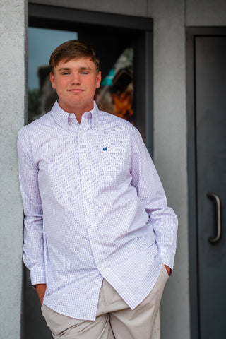 A young man wearing a light purple and white gingham long-sleeve button-down shirt with beige pants, standing in front of a gray building wall.