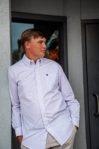 A young man wearing a light blue and white micro-check button-down shirt with beige pants, standing in front of a gray concrete wall and doorway.