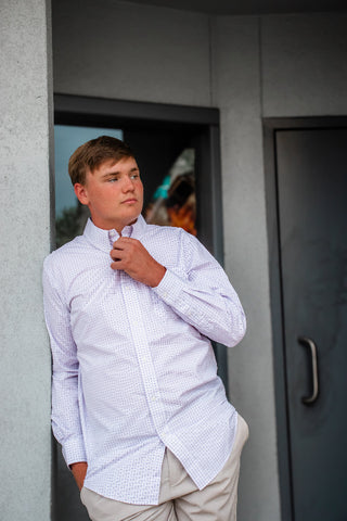A young man wearing a light blue and white micro-check patterned dress shirt and beige trousers, adjusting his collar in front of a gray concrete wall.