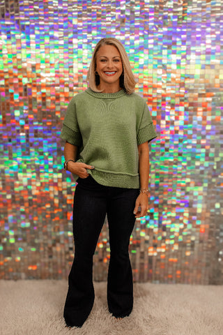 A woman stands in front of a colorful, reflective mosaic wall, wearing a sage green short-sleeve knit sweater with ribbed cuffs and hem, paired with black flared pants.