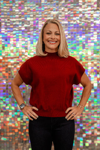 A smiling blonde woman wearing a red short-sleeve mock-neck top and dark jeans, standing with hands on hips in front of a colorful, reflective mosaic wall.