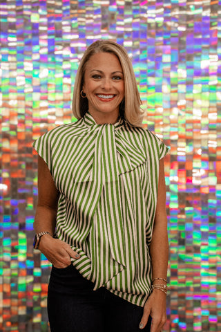 A smiling blonde woman wearing a green and white vertically striped sleeveless blouse with a ruffled tie-neck detail, posed in front of a colorful, reflective mosaic tile wall.
