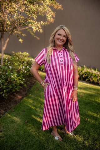 A woman stands on a grassy lawn wearing a pink and white vertically striped short-sleeve midi dress with a button-down collar. The background features a brown stucco wall and green bushes under warm sunlight.