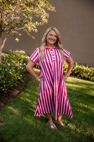 A woman stands outdoors on a grassy lawn wearing a pink and white vertically striped short-sleeve collared dress with a button-front placket and a flared skirt.