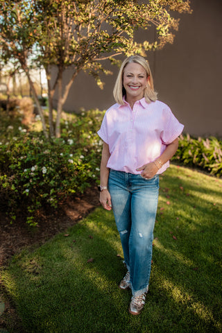 A woman stands on a grassy lawn wearing a light pink short-sleeve button-up shirt, blue jeans, and patterned sneakers, with trees and shrubs in the background.