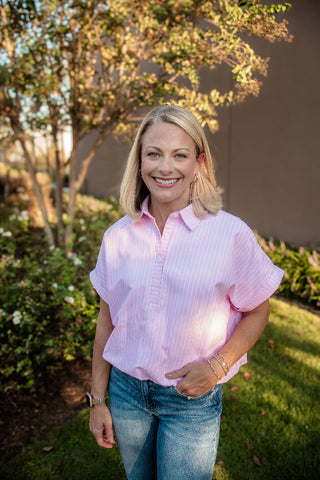 A smiling blonde woman wearing a light pink and white vertically striped short-sleeve button-up shirt and blue jeans, standing outdoors in a garden setting with trees and grass in the background.
