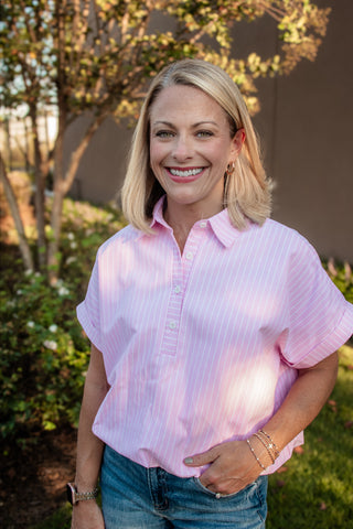 A smiling blonde woman wearing a light pink short-sleeve button-up shirt with thin white vertical stripes and blue jeans, standing outdoors in front of trees and a building wall.