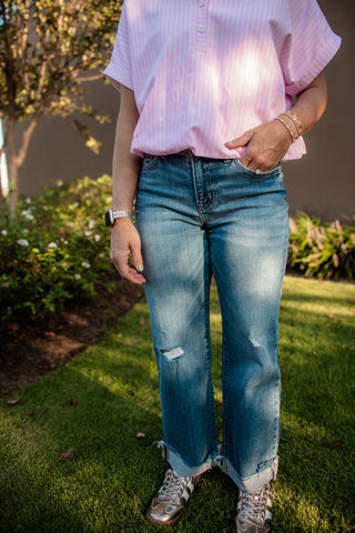 A person standing outdoors on a grassy lawn wearing light-wash blue distressed jeans with rolled cuffs, a light pink striped short-sleeve button-up shirt, and metallic sneakers.