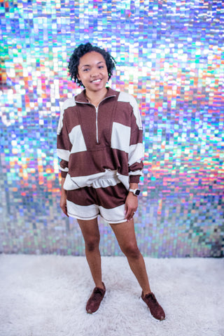 A woman modeling a matching brown and white color-blocked lounge set consisting of a quarter-zip sweatshirt and shorts, paired with brown loafers, standing in front of a colorful holographic mirror background.