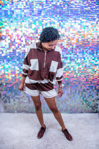 A woman wearing a brown and white color-blocked half-zip sweatshirt and matching shorts, standing on a white shag rug in front of a colorful holographic mirror backdrop.
