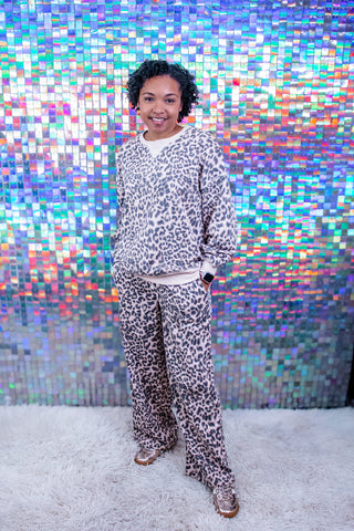 A person wearing a matching beige and black leopard print sweatshirt and sweatpants set, standing in front of a colorful holographic tile wall on a white shag rug.