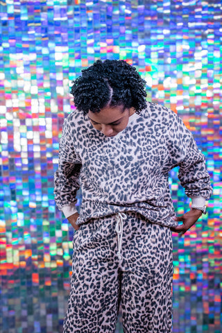 A person wearing a matching leopard-print fleece lounge set, looking down, set against a vibrant, colorful holographic tile background.