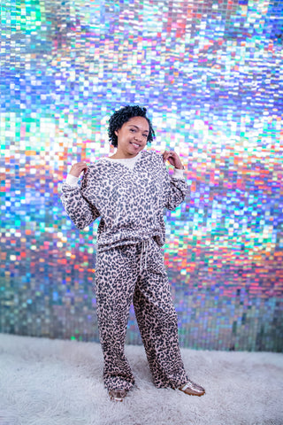 A person modeling a matching two-piece lounge set with a beige and black leopard print pattern, standing in front of a colorful, iridescent pixelated backdrop.