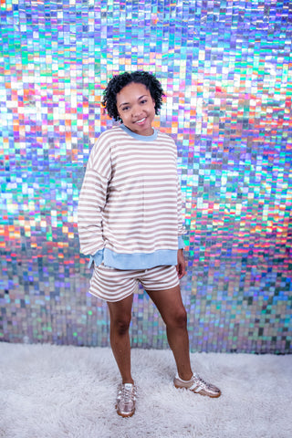A woman modeling a matching beige and white striped lounge set with a light blue hem detail, standing on a white shag rug in front of a colorful holographic pixelated backdrop.