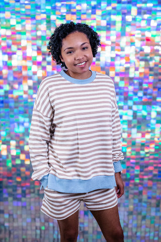 A woman modeling a matching beige and light blue striped lounge set, consisting of a long-sleeve sweatshirt and shorts, posed in front of a colorful, reflective mosaic tile wall.