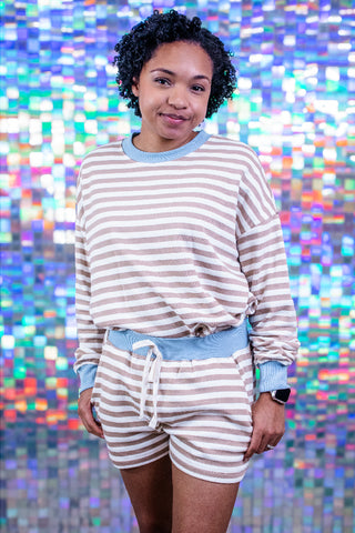 A woman modeling a matching beige and light blue striped lounge set, consisting of a long-sleeve shirt and shorts with a drawstring waist, posed against a colorful, reflective holographic background.