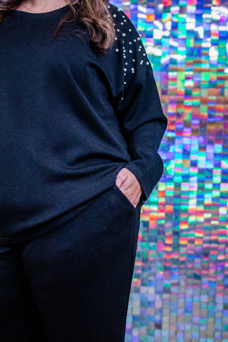 A person wearing a dark navy long-sleeve top with silver rhinestone embellishments on the shoulder, paired with matching dark pants, standing in front of a colorful holographic mosaic wall.