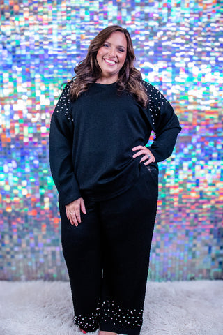 A woman modeling a black two-piece lounge set with silver rhinestone embellishments on the shoulders and cuffs, standing in front of a colorful, reflective pixelated backdrop.