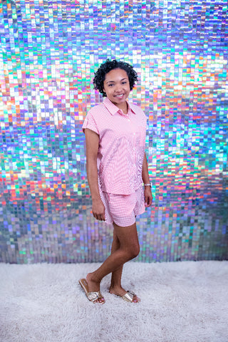 A woman modeling a pink and white striped short-sleeve button-up shirt and matching shorts set, standing on a white shag rug in front of a colorful holographic tile wall.