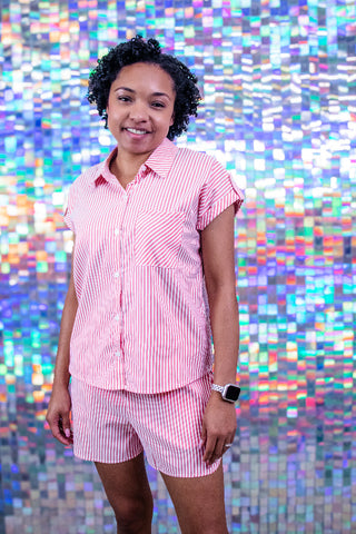 A woman wearing a pink and white vertically striped short-sleeve button-up shirt and matching striped shorts, standing in front of a colorful, reflective mosaic tile wall.