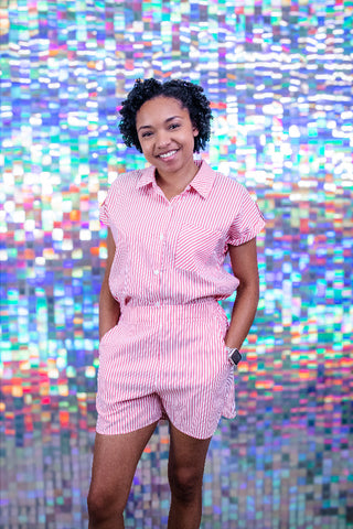 A woman wearing a pink and white striped short-sleeve button-up shirt and matching striped shorts, standing in front of a colorful, pixelated mosaic background.