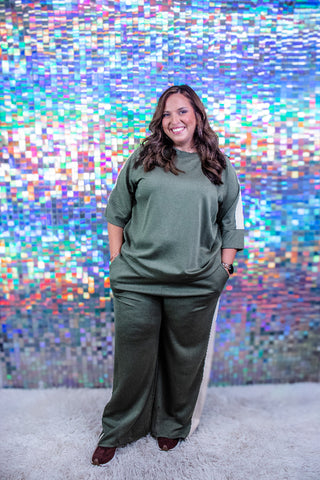 A woman wearing a matching olive green lounge set with a short-sleeve top and wide-leg pants, standing in front of a colorful, reflective mosaic tile wall.