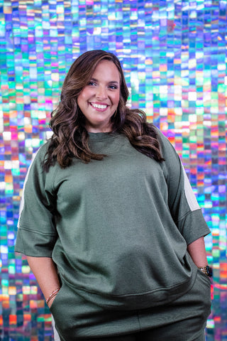 A woman with long brown hair smiling in front of a colorful mosaic tile wall, wearing an olive green sweatshirt with white side panels and matching green sweatpants.