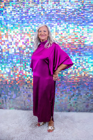 An older woman with long gray hair stands on a white shag rug in front of a colorful holographic mirror backdrop, wearing a flowing magenta-purple satin dress with draped sleeves and metallic gold sandals.