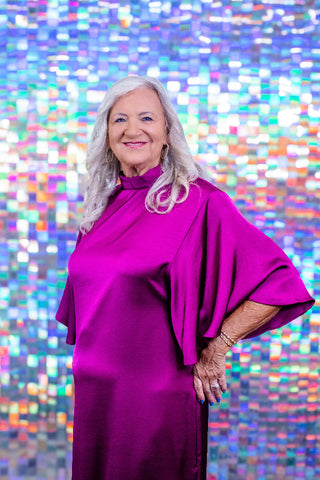 An older woman with long gray hair wearing a vibrant magenta-purple high-neck dress with wide draped sleeves, standing in front of a colorful, reflective mosaic tile wall.