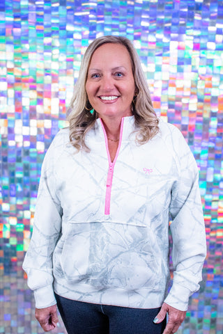 A smiling blonde woman wearing a white quarter-zip pullover with a pink zipper and a subtle gray marble pattern, standing in front of a colorful holographic tile wall.