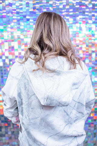 A woman viewed from behind wearing a white hoodie with a gray marble pattern, standing in front of a colorful, reflective mosaic wall.
