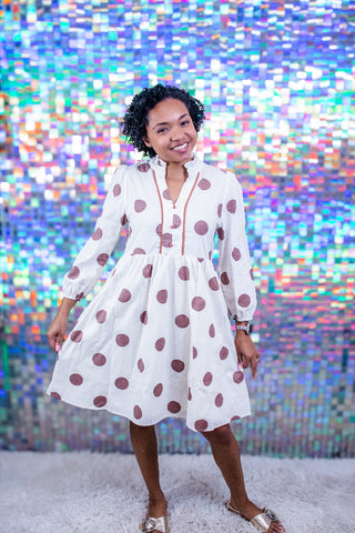 A woman modeling a white long-sleeve midi dress with brown polka dots, standing in front of a colorful, reflective mosaic wall.