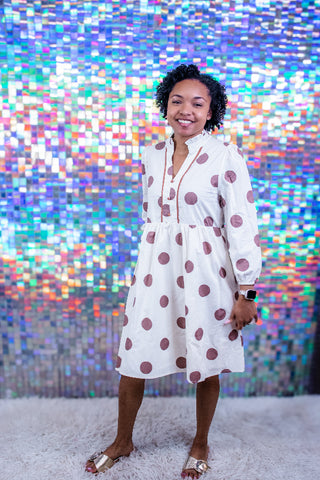 A woman wearing a white long-sleeve midi dress with large dusty rose polka dots, standing in front of a colorful, reflective pixelated backdrop on a white shag rug.