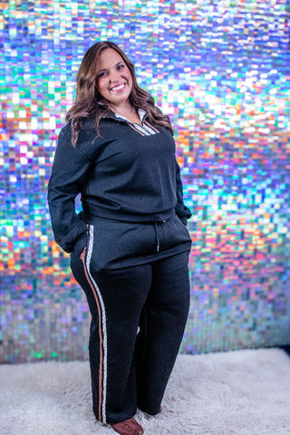 A woman modeling a black two-piece tracksuit with a striped inner collar and side stripes, standing in front of a colorful holographic backdrop.