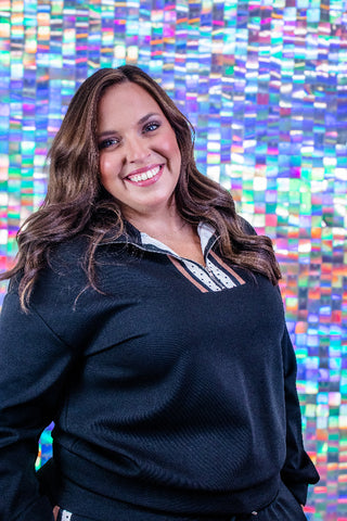 A smiling woman with long brown hair wearing a black long-sleeve quarter-zip pullover with a patterned collar, standing in front of a colorful mosaic tile wall.