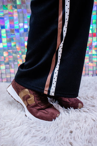 A person's legs wearing black track pants with a vertical white stripe and a metallic rose gold stripe, paired with burgundy suede sneakers with gold accents, standing on a white shag rug in front of a colorful mirrored background.