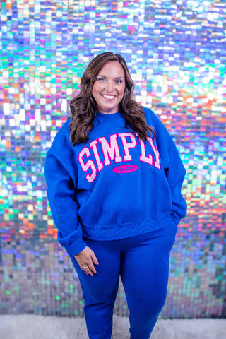 A woman wearing a bright blue sweatshirt and matching sweatpants set, featuring the word 'SIMPLY' in pink varsity lettering on the chest, standing in front of a colorful, reflective mosaic wall.