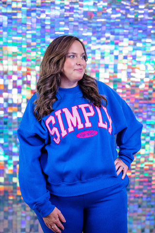 A woman wearing a bright blue sweatshirt and matching sweatpants stands in front of a colorful, reflective mosaic background. The sweatshirt features the word 'SIMPLY' in large pink letters outlined in white, with a small pink oval logo underneath.