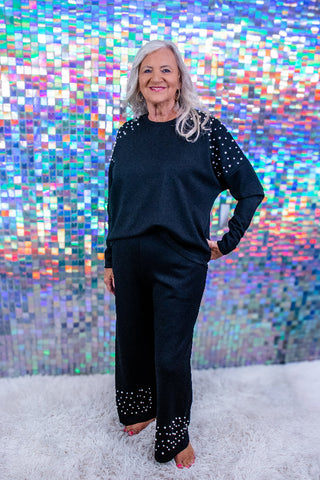 A woman with gray hair stands barefoot on a white shag rug in front of a colorful holographic mosaic tile wall, wearing a matching black lounge set with silver rhinestone embellishments on the sleeves and pant cuffs.